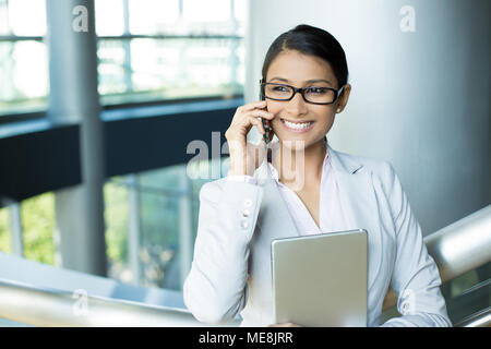 Closeup portrait, attractive happy woman in gray white suit and black glasses using silver pc and phone, isolated indoors interior office background.  Stock Photo