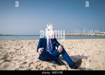 Young man in funny mask sits on the sand and enjoys vacation. Unusual guy in stylish suit relaxes on the beach. Unicorn on background of sea and sky Stock Photo