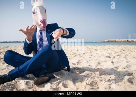 Stylish man in funny mask and elegant suit sits on beach and looks on the watch. Scared man lost in time and is late for business Stock Photo