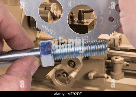 Measuring by screw pitch gauge with lathe in background. Measurement of thread on steel bolt with nut and detail of turner's hands in workplace. Stock Photo