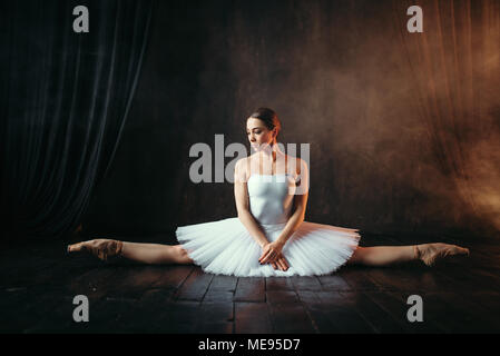 Ballerina in white dress sits on a twine, front view. Body flexibility of classical ballet dancer