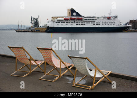 Deckchairs left on the quay in gray and cold weather. Facing a cruise ship, symbolizing a longing for summer and travel. Dreaming of travel and vacati Stock Photo