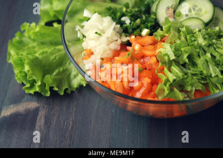 Glass bowl with cut vegetables for a salad. Close up. Bell pepper, onion, leafy greens and cucumbers cutted in pieces. Wooden background. Stock Photo