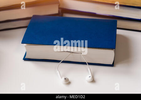 The book lies on a white table against the background of old yellowed books. Headphones stick out from the book. New technologies have replaced the ol Stock Photo