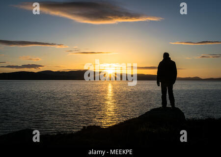 This is a silhouette of a person watching the sun go down behind some mountains at the sea.  This photo was taken near Buncrana Ireland Stock Photo