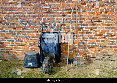 Traditional gardening tools wheelbarrow two rakes a fork and a plastic flexi tub by an old brick wall  in Helmsley Walled Garden North Yorkshire Stock Photo