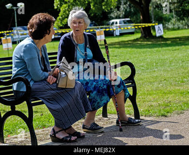 Two elderly women sit on a bench talking at Canons Park, Harrow, London. One holds a flowered walking cane. Stock Photo