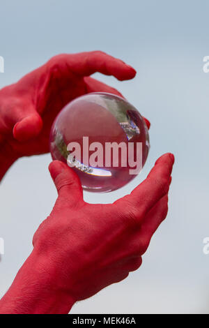 man with red hands dressed as the devil holds and plays with an isolated glass crystal ball containing reflections of people Stock Photo