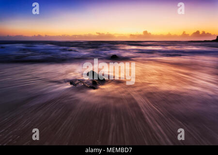 Warm sunlit sunrise at sandy beach in Nambucca heads regional town of Australia - pacific coast. Smooth lines of waves draw around rocks in gentle tid Stock Photo