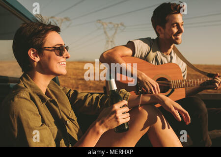 Couple sitting in their car enjoying the road trip in country side. Man playing the guitar while the woman is holding a bottle of drink. Stock Photo