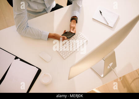Top view of a man using mobile phone sitting in front of computer with documents and coffee glass on the table. Man using wireless keyboard and mouse  Stock Photo