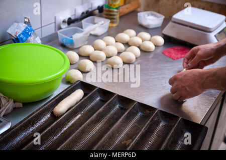A close-up of a female bakerpulls out the dough for cooking buns for hotdogs on a metal  becary table with a rolling pin Stock Photo