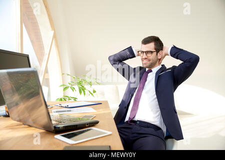 Portrait of young businessman in suit and glasses using laptop while sitting with hands on his hand and working on new project. Stock Photo