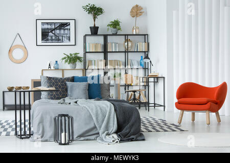 Orange armchair next to grey bed in bedroom interior with poster on white wall above bookshelf Stock Photo