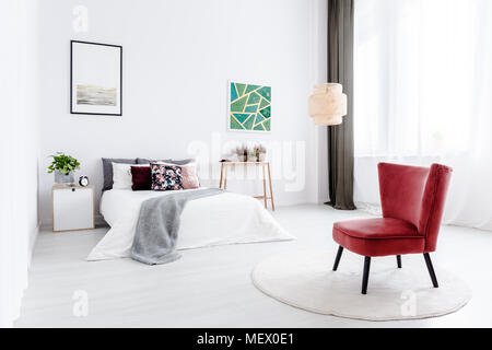 Red armchair on round rug in white bedroom interior with green painting and poster on the wall Stock Photo