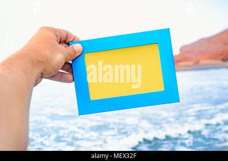 A male hand holds a blue photo frame with empty space seascape on background. Travel concept. Stock Photo