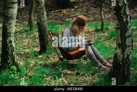 young caucasian female  artist sitting outdoors in a forest and drawing a picutre into her notebook Stock Photo