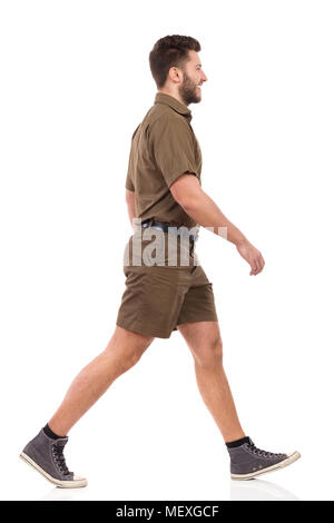 Male Messenger In Khaki Uniform Is Walking, Smiling And Looking Away. Side View. Full Length Studio Shot Isolated On White. Stock Photo