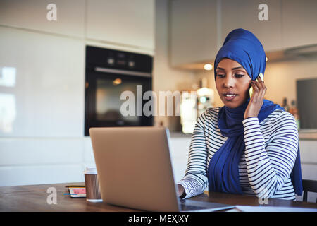 Young Muslim female entrepreneur wearing a hijab talking on a cellphone and working on a laptop while sitting at a table in her kitchen Stock Photo