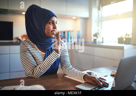 Young Arabic female entrepreneur wearing a hijab looking deep in thought while sitting at her kitchen table working on a laptop Stock Photo