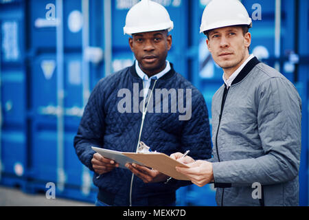 Two engineers tracking inventory using a clipboard and digital tablet while standing by freight containers in a large commercial shipping yard Stock Photo
