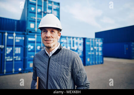 Port manager wearing a hardhat and smiling while standing alone on a large commercial shipping dock holding a clipboard Stock Photo