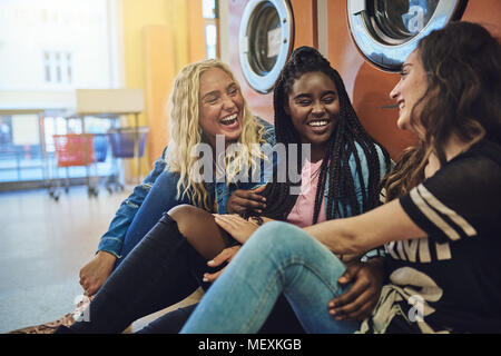 Diverse group of young girlfriends sitting together on a laundromat floor laughing and talking while doing laundry Stock Photo