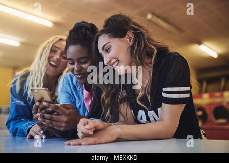 Laughing young girlfriends leaning on a laundromat counter using a cellphone together while doing laundry Stock Photo