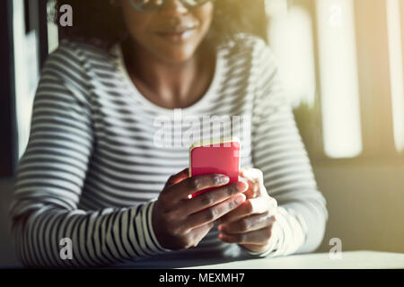 Smiling young African woman wearing glasses sitting at a table reading a text message on her cellphone Stock Photo