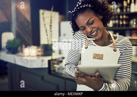 Smiling young African entrepreneur standing at the counter of her cafe talking on a cellphone and using a tablet Stock Photo