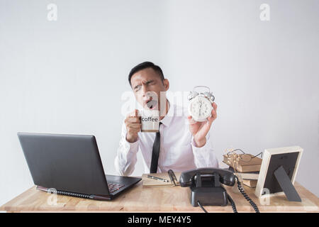 Young asian businessman wearing white shirt with tie yawning while and show alarm clock on afternoon time drinking coffee together Stock Photo