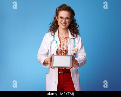 smiling paediatrician doctor in white medical robe showing tablet PC blank screen isolated on blue Stock Photo