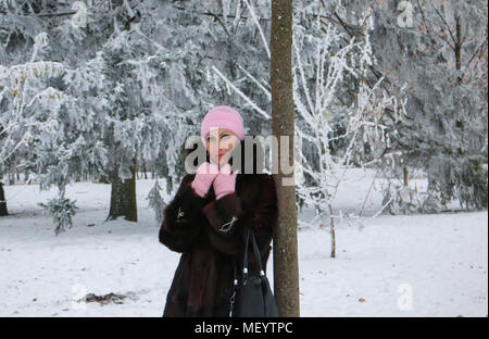 Pretty and calm woman in fur coat posing near the tree in winter snow-covered park. Cold atmosphere in the day. Forest after the snowfall. Stock Photo