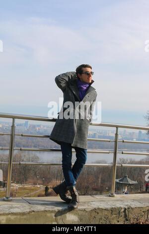 The portrait of a handsome smiling young man in the black sunglasses, who is standing on the bridge in a park. Sunny weather. Stock Photo