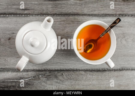 Table top view on small white teapot and cup full of hot amber tea with silver spoon in it, placed on gray wood table. Stock Photo