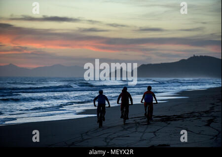 Three mountain bikers ride along a beach near the port of Punta Ala in Tuscany, Italy at sunset. Stock Photo