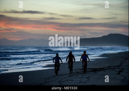 Three mountain bikers ride along a beach near the port of Punta Ala in Tuscany, Italy at sunset. Stock Photo