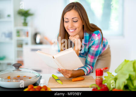 Beautiful young woman cooking healthy meal in the domestic kitchen. She is reading recipes from the cookbook. Stock Photo