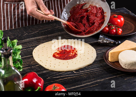 A male hand spreading tomato puree on a pizza base with spoon on an old wooden background Stock Photo