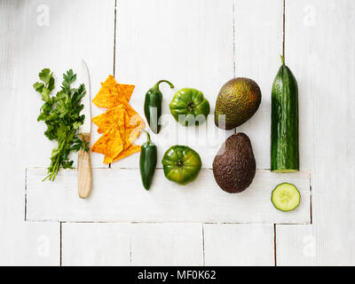 Avocados, green tomatoes, Jalapeno peppers, cucumber, parsley, kitchen knife and tortilla chips on white wood Stock Photo
