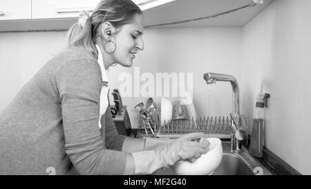 Black and white portrait of smiling young housewife washing dishes and listening music with earphones Stock Photo