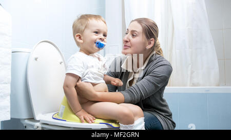 Cute smiling toddler boy sitting on toilet with young mother Stock Photo