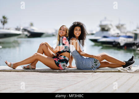 Spain, Andalusia, Marbella. Two multiracial traveler women sitting in boats harbor. Lifestyle concept. Stock Photo