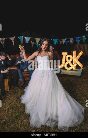 Happy bride holding cocktail glass on a night field party with groom and friends in the background Stock Photo