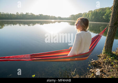 Young woman relaxing on hammock by the lake, sunlight sunbeam and reflection. People relaxation wellbeing in nature concept. France, Europe Stock Photo