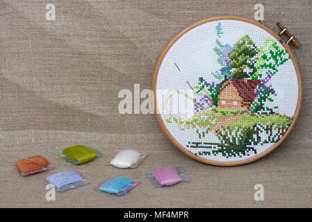 The cross-stitched pattern of a house is in an upright position on a grey linen background. The bobbins of color mouline are next to a Stock Photo