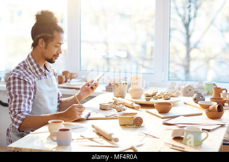 Young mixed race craftsman sitting at table with handmade clay pots, using smartphone and checking orders in notebook Stock Photo