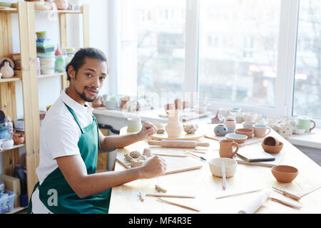 Portrait of young African American male potter sitting at messy table with handicrafts and looking at camera Stock Photo