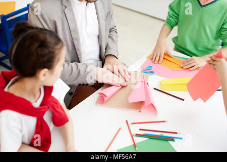 Children making paper airplanes with colored paper during art lesson while sitting at classroom desk with teacher Stock Photo