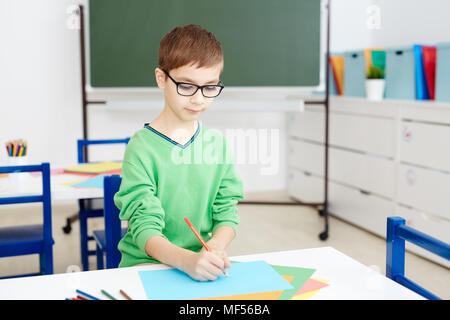 Handsome Caucasian boy in glasses sitting at classroom desk and diligently drawing on colored paper with pencil Stock Photo
